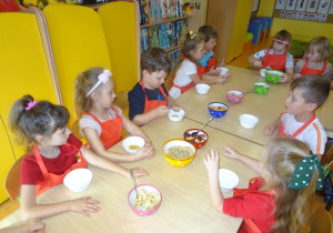 Grupa dzieci siedzi przy stole, przed nimi stoją miseczki. Dzieci łyżkami nakładają z misek rozstawionych na środku stole jabłka, mandarynki, banany, płatki.